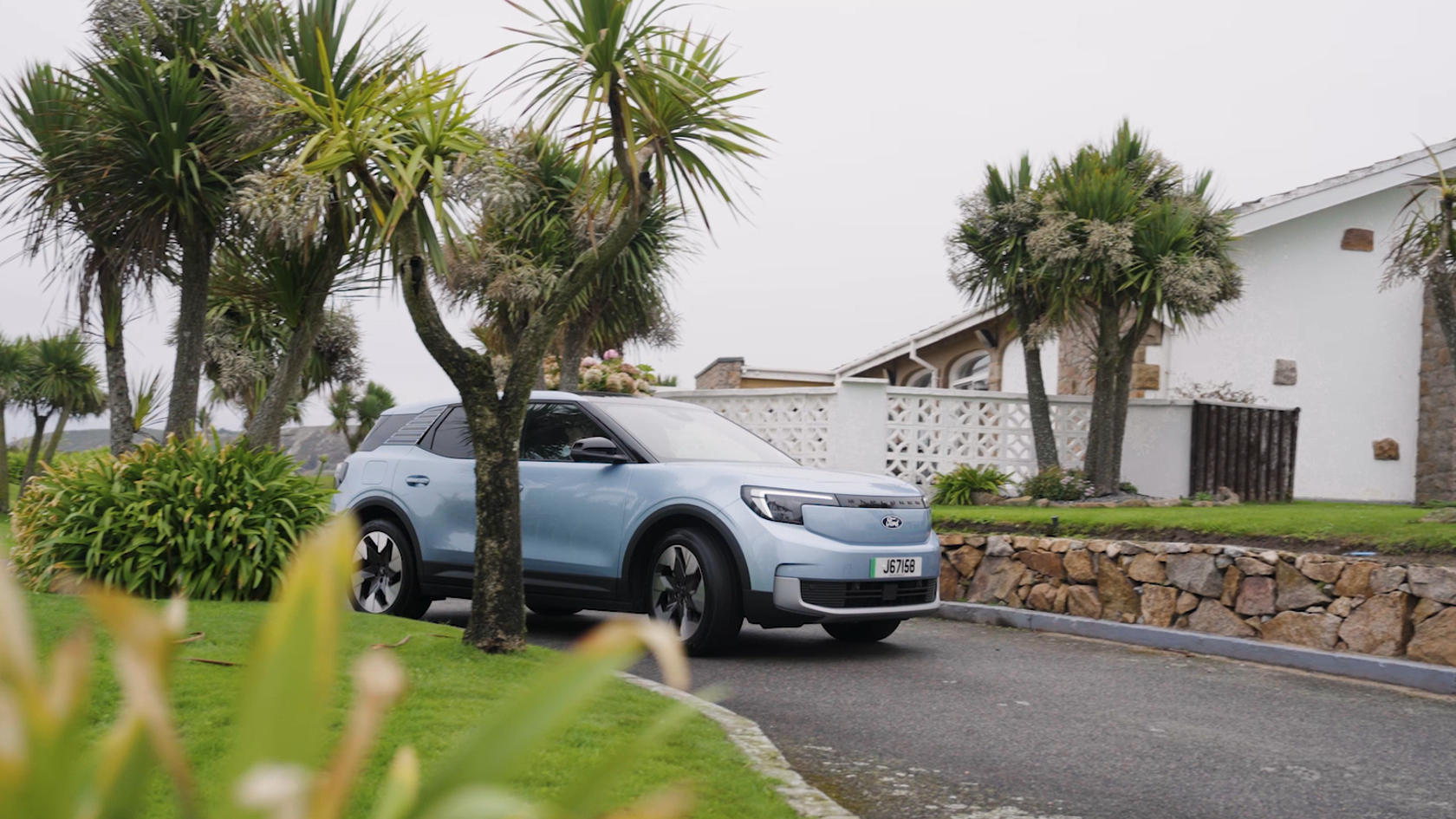 A light blue electice Ford Explorer pulling into a drivewhich has grass and palm trees either side. A house sits in the backgroud